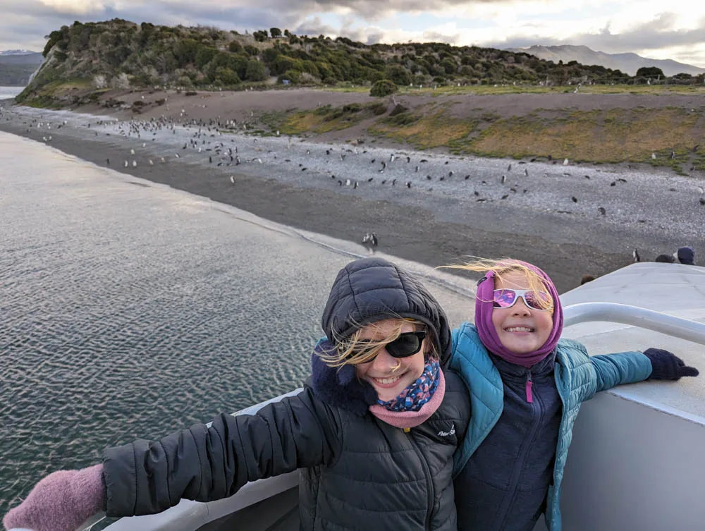 Georgia and Eva wrapped up warm on the deck of a boat with a large colony of Magellanic penguins on a beach in the background.