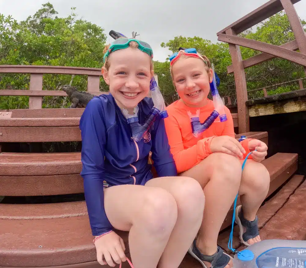 A smiling Georgia and Eva sat on steps in their snorkeling gear with a marine iguana lurking in the background.