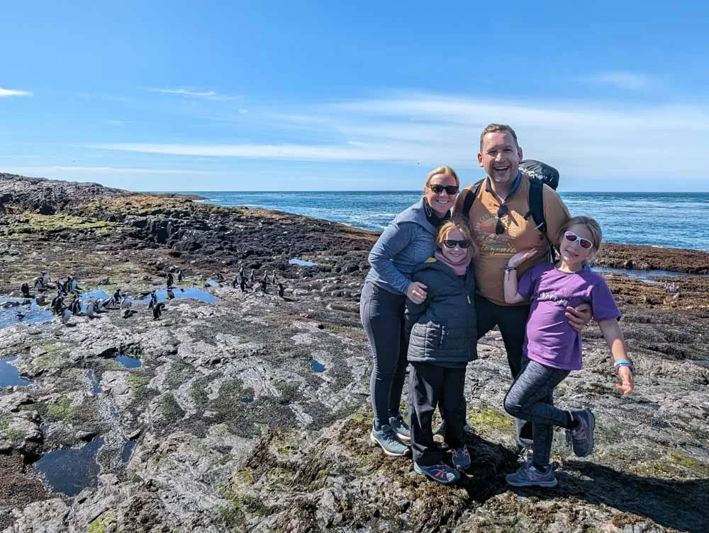 A family photo on the rocks at Penguin Island with a group of Magellanic penguins off to the left with the sea in the background and blue skies.