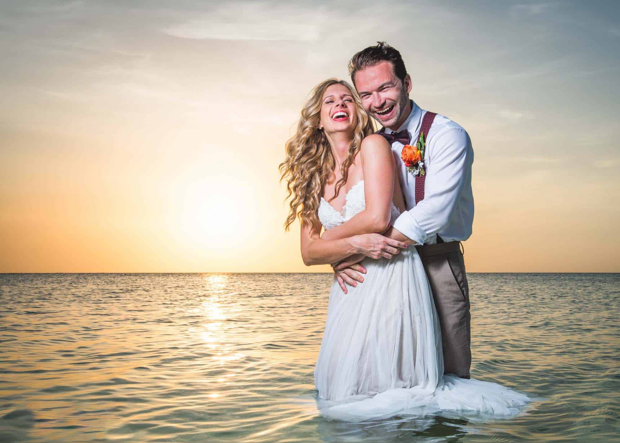 Bride and groom standing in the ocean at sunset, laughing and embracing, a stunning moment captured by Will Hey Photography.