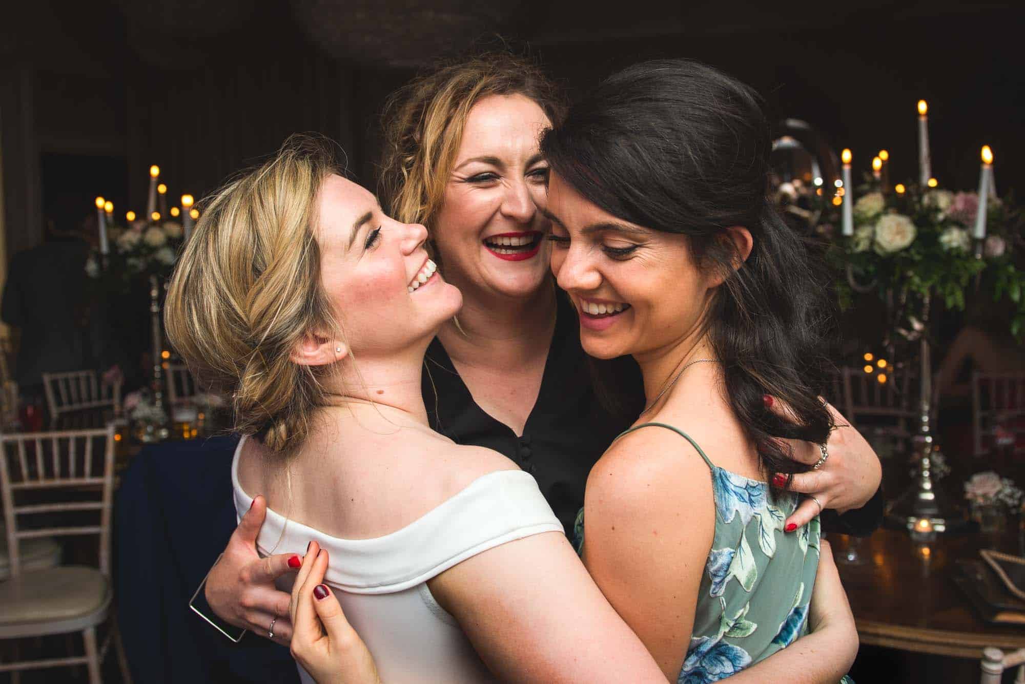 Three women laughing and hugging in a warmly lit wedding reception, their happiness captured by Will Hey Photography.