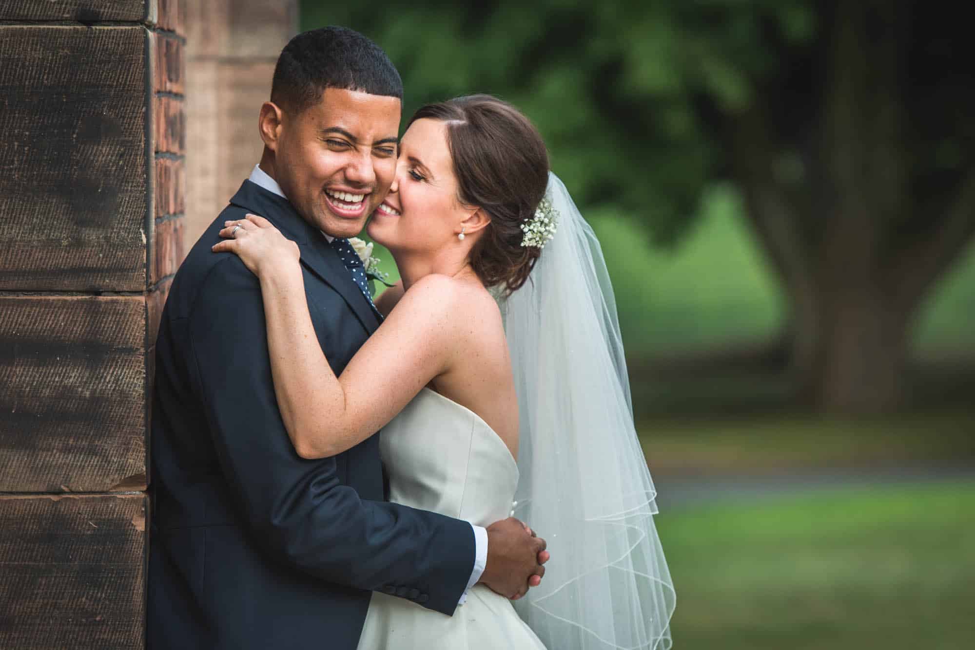 Bride and groom laughing and embracing next to a rustic wooden wall at an outdoor wedding, captured by Will Hey Photography.