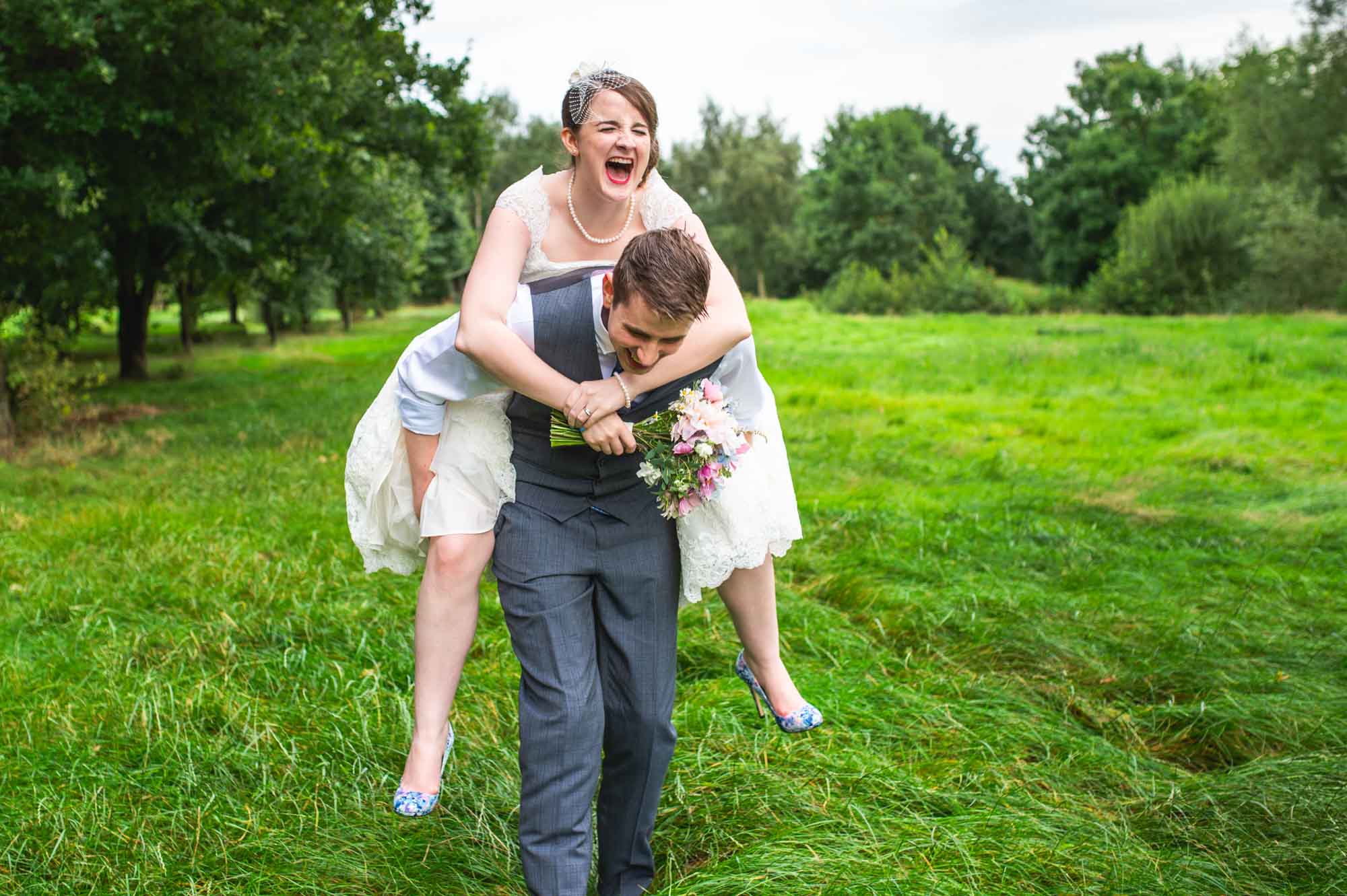 Bride laughing while being carried on the groom's back through a lush green field, holding a bouquet, taken by Will Hey Photography.